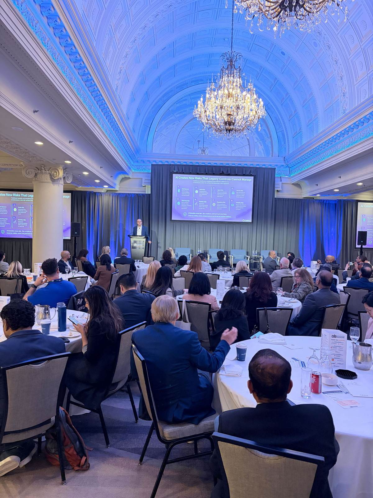 Thomas Mazzaferro speaking from a lectern in a ballroom, with a slide behind him on AI risks that legacy controls cannot manage.