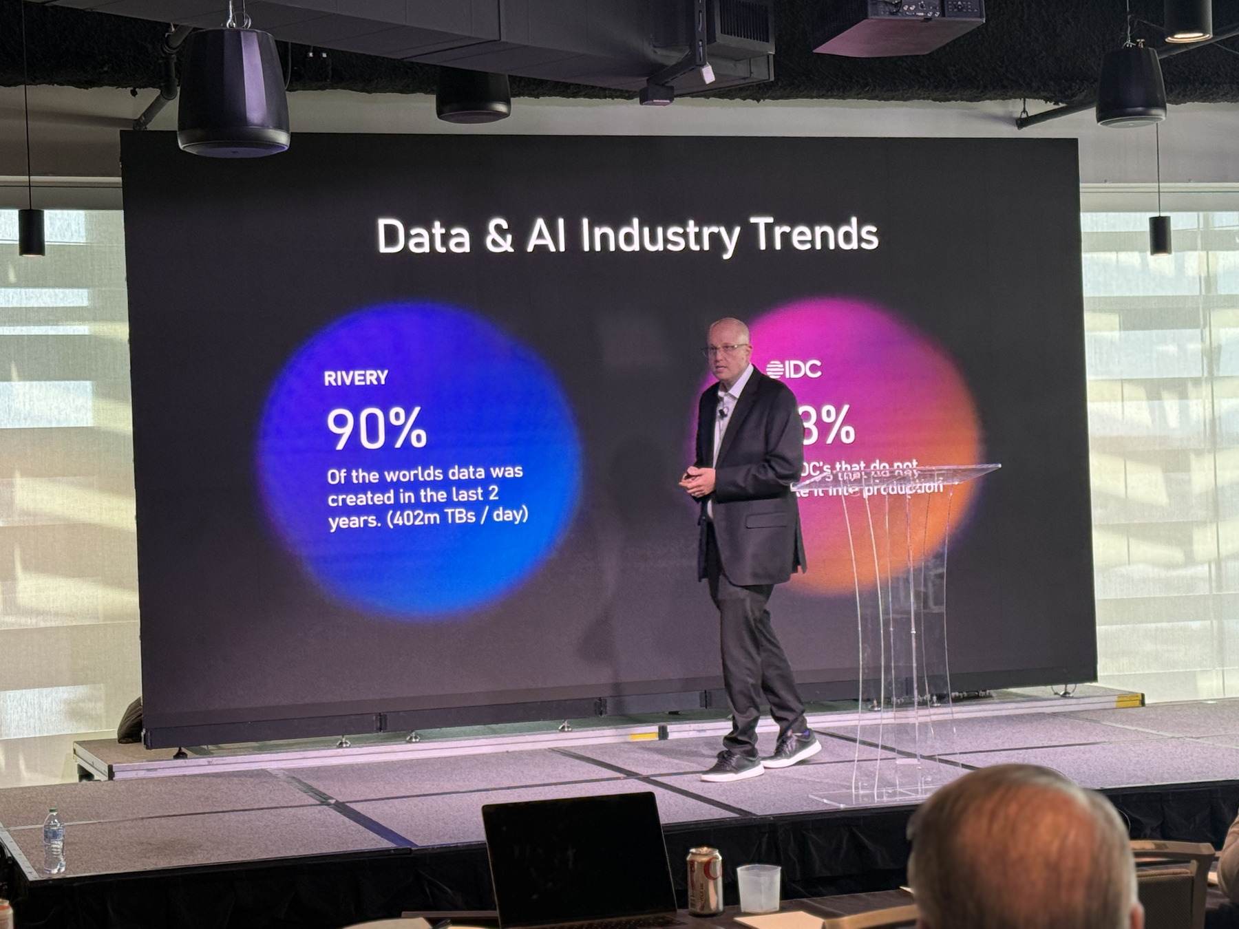 Thomas Mazzaferro speaking on stage in front of a large screen showing data and AI industry trend statistics.