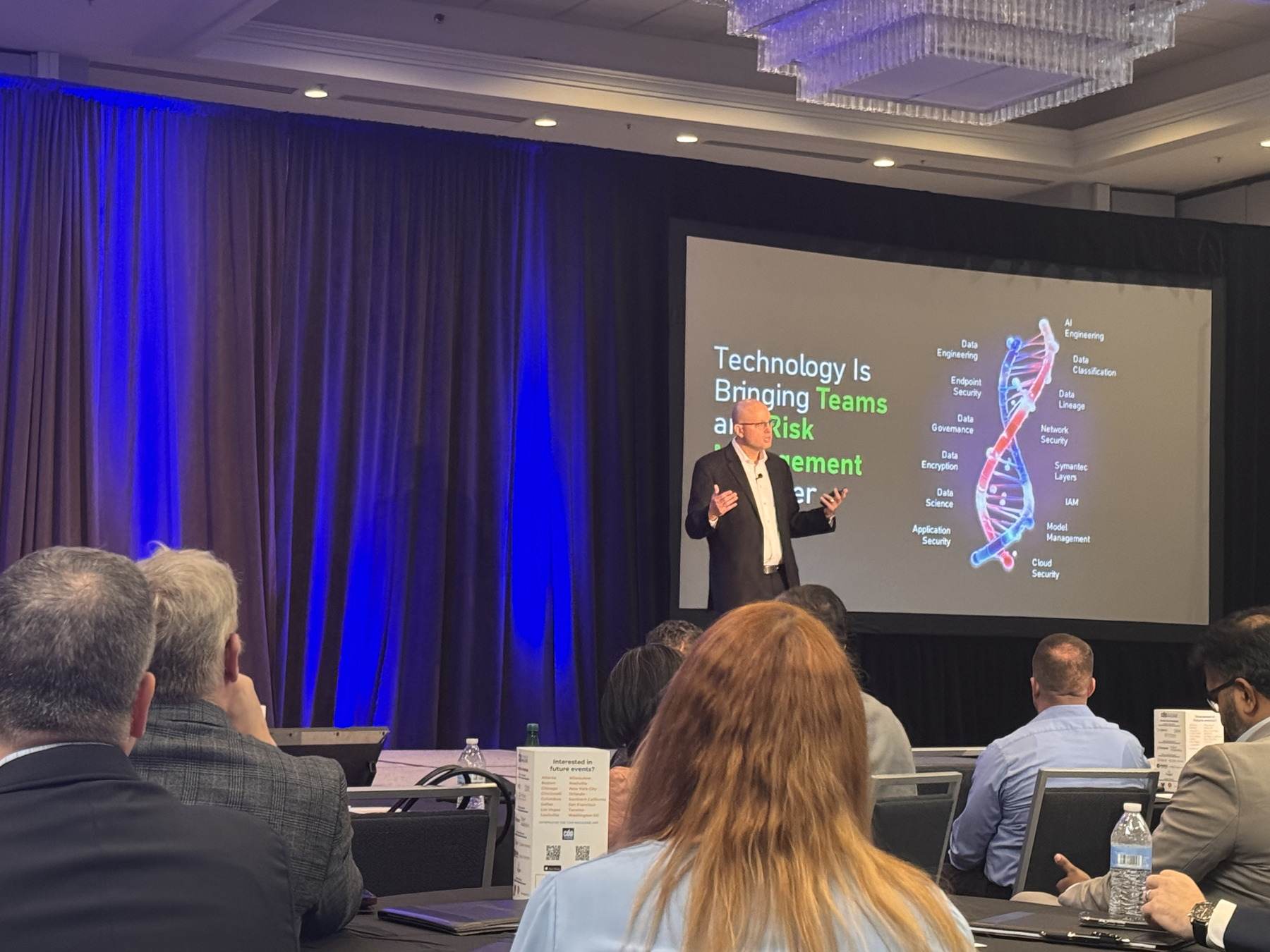 Thomas Mazzaferro presenting to a seated audience beside a slide showing how technology brings teams and risk management together.